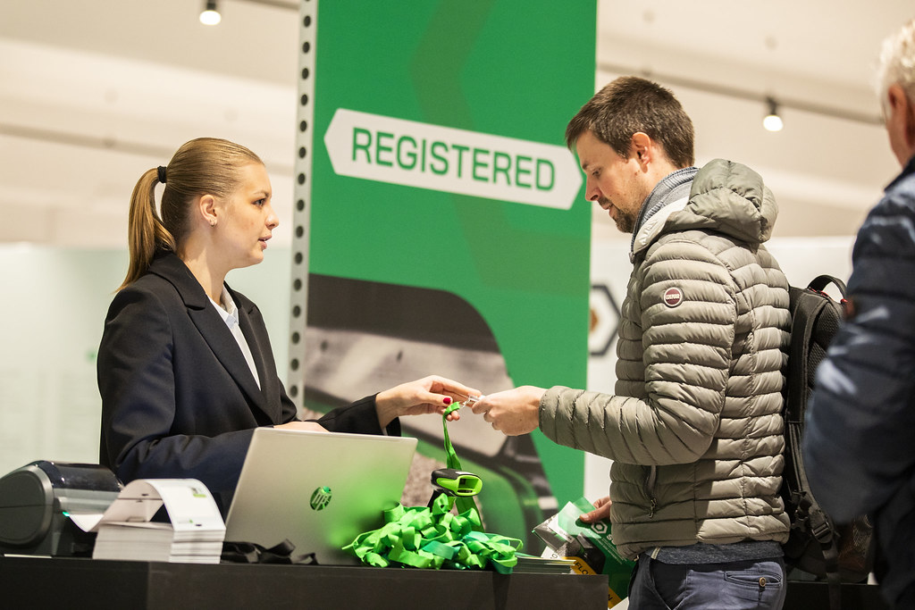 Hostess overhandigt een badge aan een bezoeker bij de registratiebalie onder een groen REGISTERED-bord