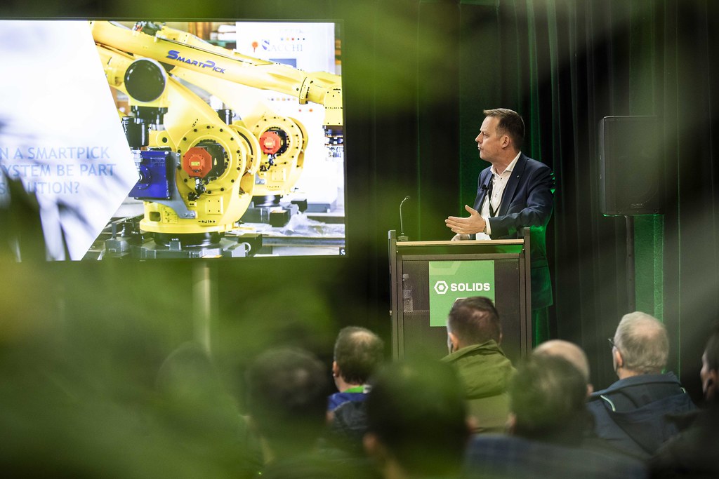 Spreker achter een SOLIDS-spreekgestoelte voor een zaal met toehoorders, met een presentatie over robotsystemen op het scherm