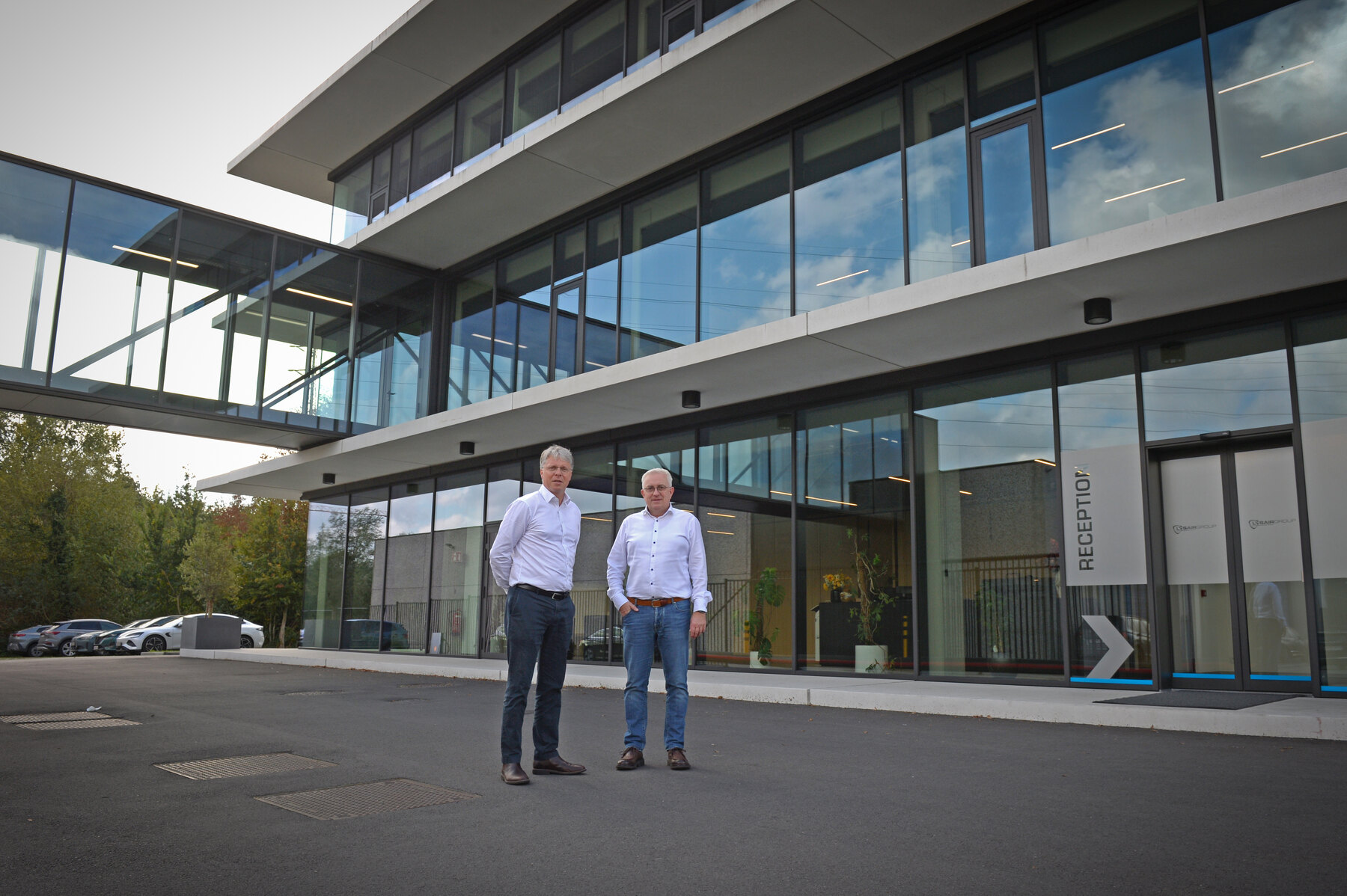 Alex Vanwolleghem en Herbert Vandaele staan in witte overhemden voor het glazen kantoorgebouw van Opina (voorheen LysairGroup) in Sint-Baafs-Vijve
